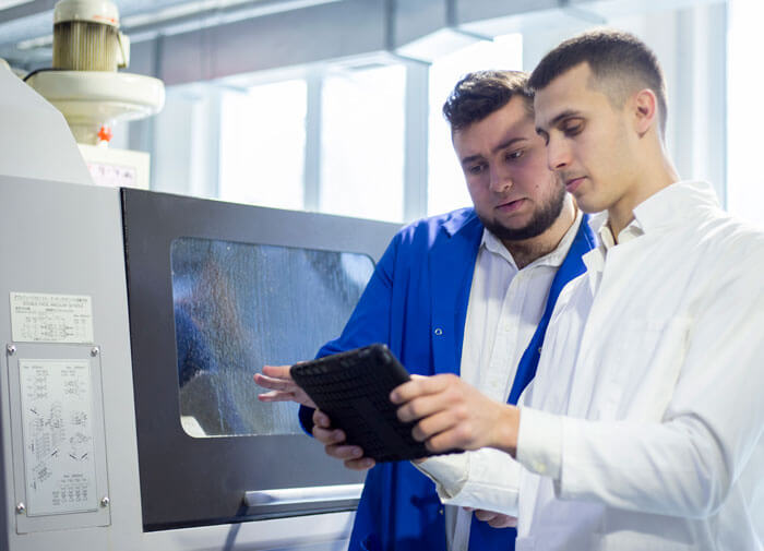 Zwei Männer, einer im weißen Kittel, einer in blauer Arbeitskleidung, betrachten ein Tablet vor einer Industriemaschine.